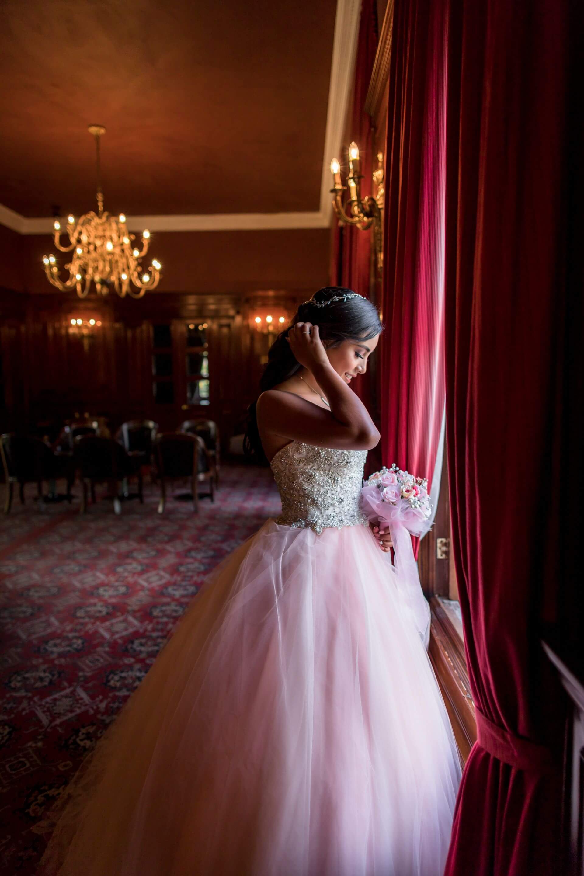 A quinceañera in a pink dress standing beside a sunlit window